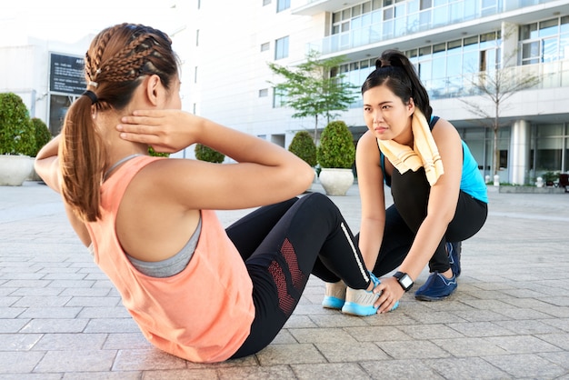 Ajuste mujer asiática haciendo sentadillas en el pavimento en la calle y amiga sosteniendo sus pies