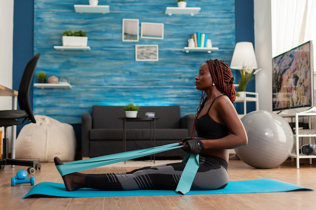 Ajuste la sala de estar en casa de entrenamiento de mujer afro fuerte usando una banda de resistencia, sentado en una colchoneta de ejercicios tirando de los músculos de la espalda