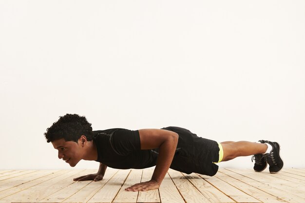 Ajuste sonriente joven atleta negro haciendo flexiones sobre un piso de madera clara contra una pared blanca