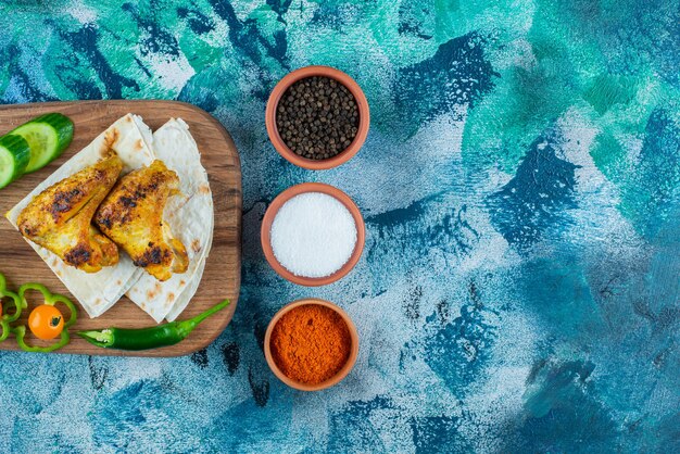 Alas cocidas, lavash y verduras sobre una tabla de cortar, sobre fondo azul.