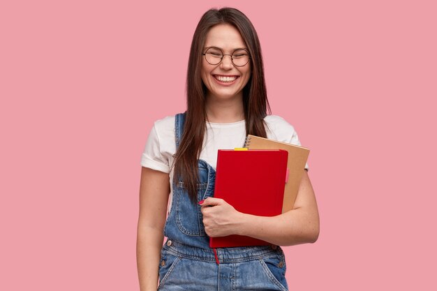 Alegre adolescente sonriente con expresión complacida, lleva dos libros de texto, tiene una gran sonrisa, se ríe de algo bueno