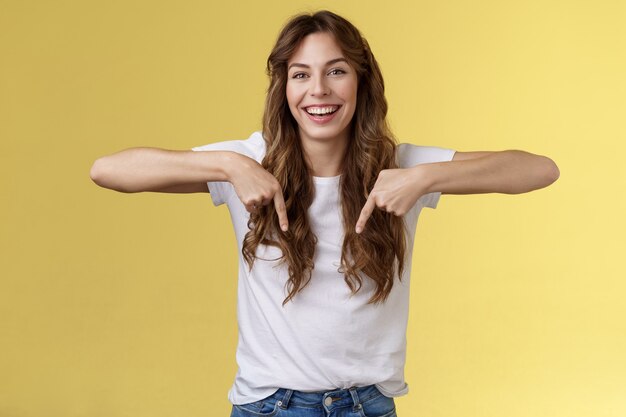 Alegre alegre alegre niña hermosa cabello largo apuntando hacia abajo los dedos índices indican promoción inferior riendo alegremente compartir emociones positivas recomendar publicidad de productos fondo amarillo