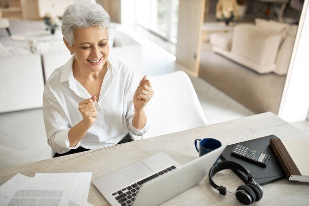 Alegre, alegre, elegante, mujer madura, agente de bienes raíces, apretando los puños, expresando entusiasmo después de hacer un buen trato en línea, sonriendo ampliamente, sentado frente a una computadora portátil abierta