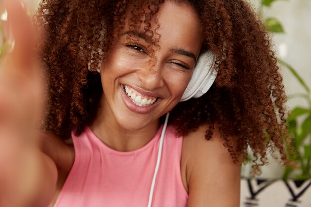 Alegre y alegre mujer africana americana posa para selfie, tiene una amplia sonrisa, escucha su pista favorita en auriculares, disfruta del tiempo de recreación, se sienta en la acogedora cafetería. Concepto de personas y entretenimiento.