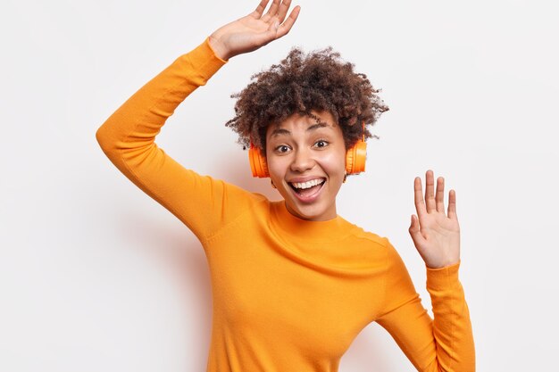 Alegre alegre mujer afroamericana disfruta de una calidad de sonido impresionante usa auriculares inalámbricos estéreo escucha bailes de música favorita con ritmo vestida con un jersey naranja aislado en una pared blanca