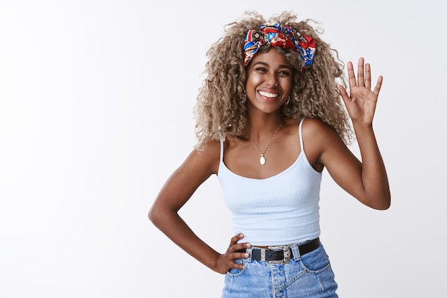 Alegre y amistosa chica afroamericana rubia con diadema hipster de peinado afro sonriendo felizmente levantando la palma de la mano y saludando en saludo hola o hola gesto ver persona familiar fondo blanco