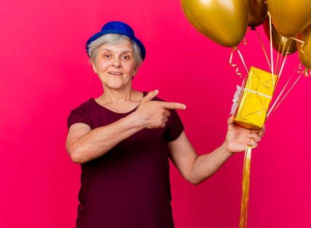 Alegre anciana con gorro de fiesta sostiene globos de helio y puntos en caja de regalo en rosa