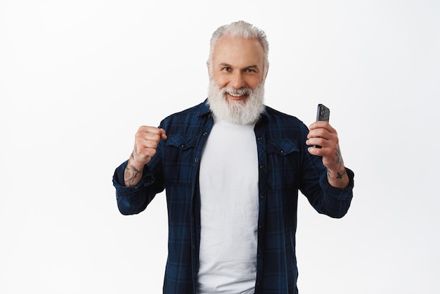 Alegre anciano ganando en el teléfono móvil celebrando y sonriendo complacido sosteniendo el teléfono inteligente y apretando los puños en triunfo lograr el objetivo de pie sobre fondo blanco