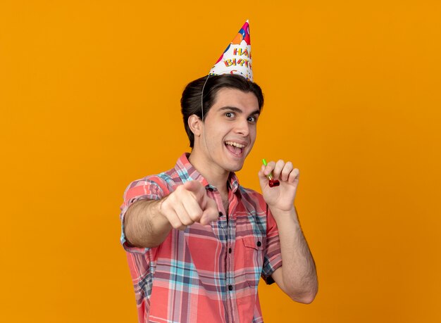 Alegre apuesto hombre caucásico vestido con gorro de cumpleaños tiene silbato de fiesta apuntando a la cámara