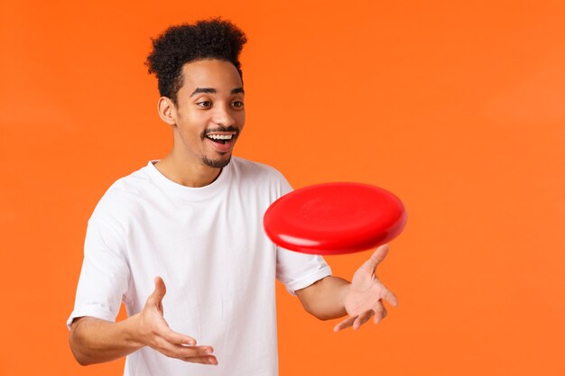 Alegre apuesto joven afroamericano saliente con corte de pelo afro, bigote, sonriendo divertido capturando frisbee rojo como jugando al aire libre, como juegos activos, fondo naranja