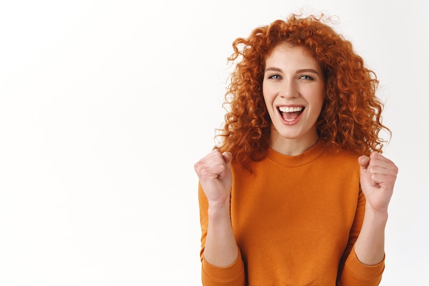 Alegre, atractiva, elegante, caucásica pelirroja apoyándote, triunfando y regocijándose de ganar, logrando la meta sonriendo optimista, puñetazo de éxito y felicidad