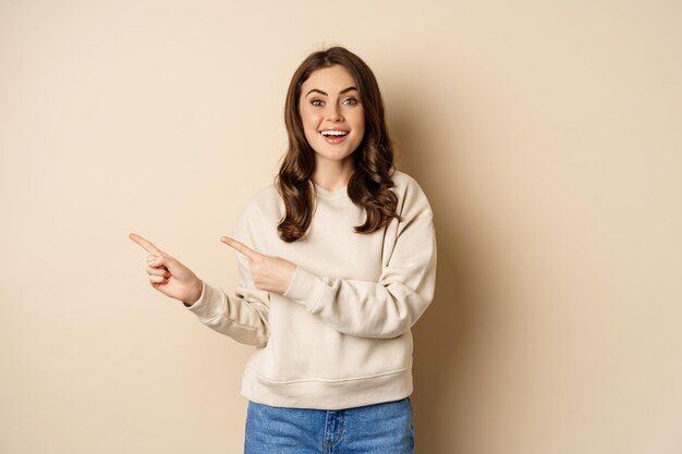 Alegre y atractiva modelo femenina señalando con el dedo a la izquierda, mostrando publicidad de la tienda, pancarta o logotipo, de pie en un suéter sobre fondo beige