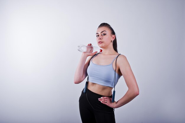 Alegre y atractiva mujer joven en fitness con top y polainas negras con cuerda para saltar y una botella de agua aislada sobre fondo blanco