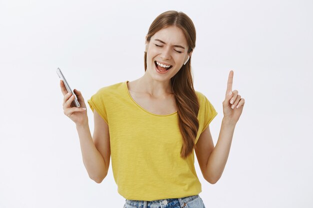 Alegre bastante joven escuchando música en auriculares inalámbricos, sosteniendo el teléfono inteligente y bailando