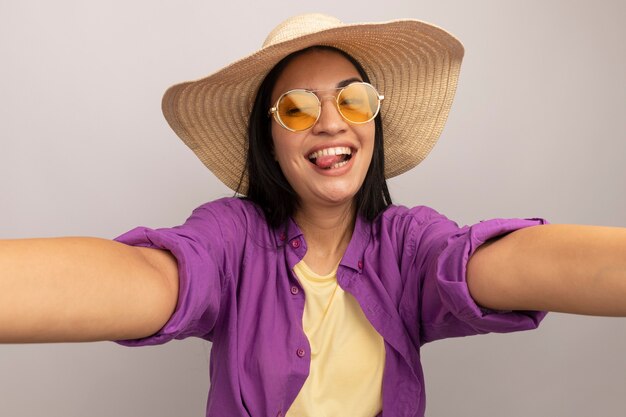 Alegre bastante morena chica caucásica en gafas de sol con sombrero de playa saca la lengua y finge sostener la cámara tomando selfie en blanco