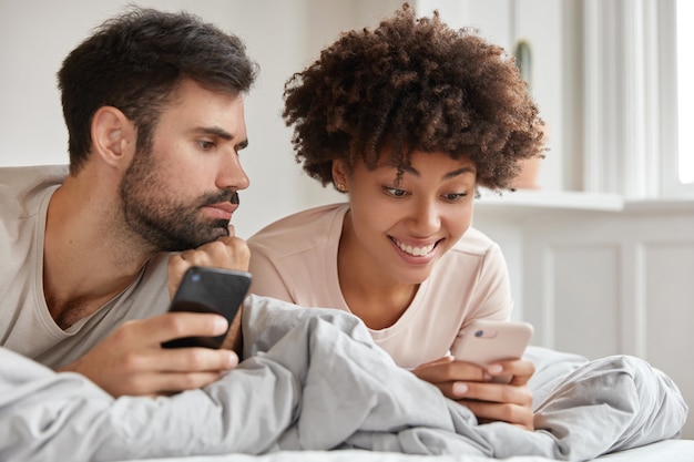 Alegre bloguera de piel oscura chatea con seguidores en teléfonos inteligentes, tiene expresión alegre, corte de pelo afro, chico sin afeitar actualiza el perfil en el celular, se acuestan juntos en la cama durante el fin de semana de la mañana