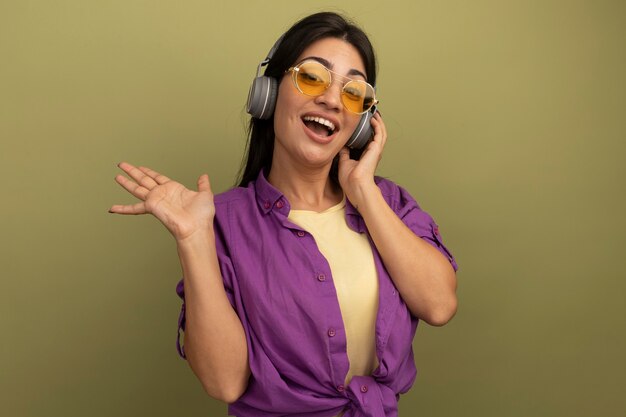 Alegre bonita morena mujer caucásica en gafas de sol se encuentra con la mano levantada aislada