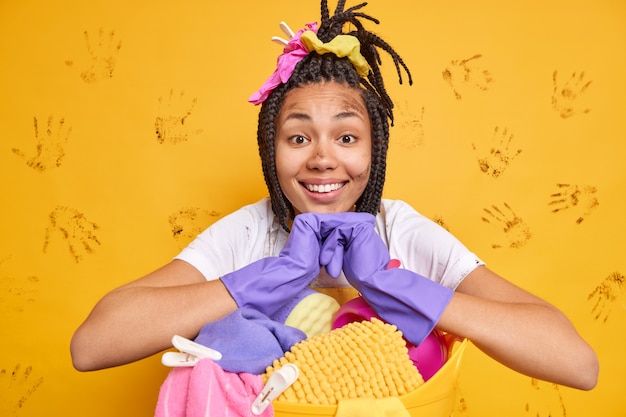 Alegre, cansada pero satisfecha mujer afroamericana usa guantes de goma se inclina en la canasta de lavandería lava todo alrededor aislado sobre pared amarilla