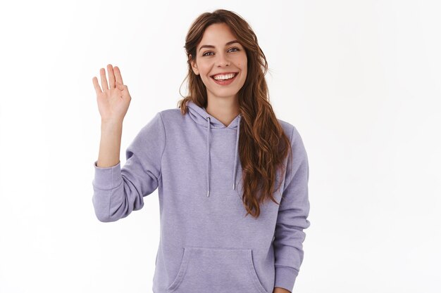 Alegre, carismática, atractiva mujer amigable agitando la mano levantada hola saludo gesto sonriente con dientes perfecta sonrisa presentarse conocer gente nueva dando la bienvenida a invitados, pared blanca