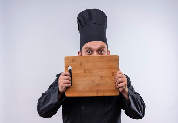 Un alegre chef barbudo hombre en uniforme negro sonriendo y mostrando tablero de cocina de madera sobre una pared blanca