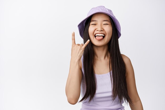 Alegre chica asiática muestra la lengua y el gesto de los cuernos de heavy metal del rock n roll disfrutando del evento divirtiéndose en el verano de pie sobre fondo blanco