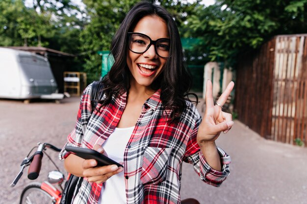 Alegre chica blanca con gafas expresando emociones positivas mientras se relaja en la ciudad. Foto al aire libre de mujer glamorosa en camisa a cuadros con smartphone.