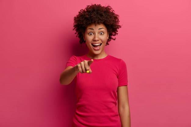 La alegre chica étnica te señala con el dedo índice, elige a alguien y sonríe alegremente, se ríe alegremente, vestida con una camiseta rosa, se para contra la pared carmesí. ¡Qué cosa tan asombrosa!