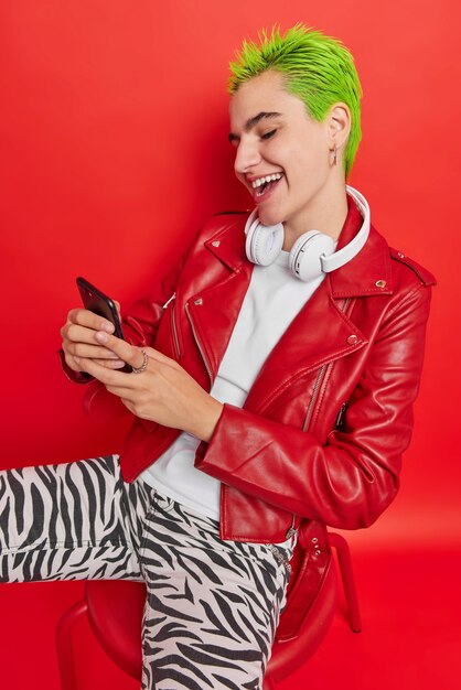 Alegre chica hipster con cabello teñido de verde usa teléfono móvil envía mensajes a amigos organiza reuniones vidas vida despreocupada disfruta de jóvenes usa una aplicación genial en el celular se sienta en una silla escucha música popular