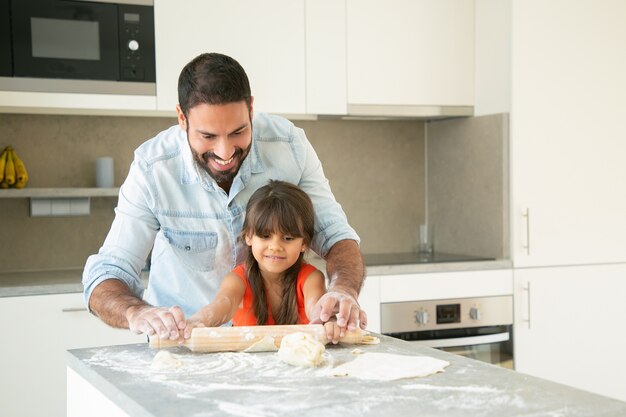 Alegre chica latina y su papá rodando y amasando masa en la mesa de la cocina con harina en polvo.