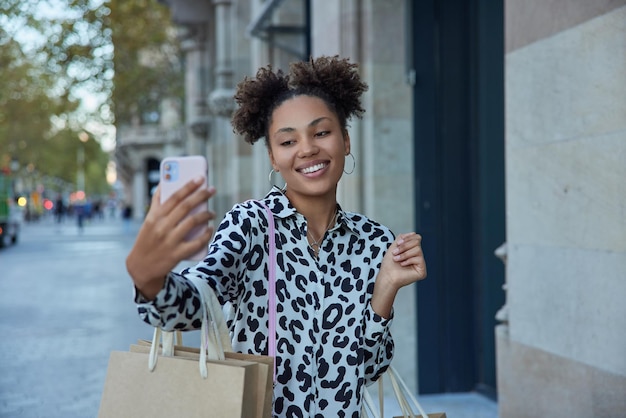 Alegre chica milenaria con cabello rizado se hace selfie a través de un teléfono inteligente moderno lleva bolsas de papel disfruta de un buen día vestida con ropa fashioable se regocija con las grandes ventas