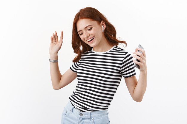 Alegre chica pelirroja con camiseta a rayas que disfruta de una calidad de sonido increíble, compró nuevos auriculares inalámbricos, sostuvo el teléfono inteligente, levantó las manos sin preocupaciones, baila y sonríe, escucha música, pared blanca