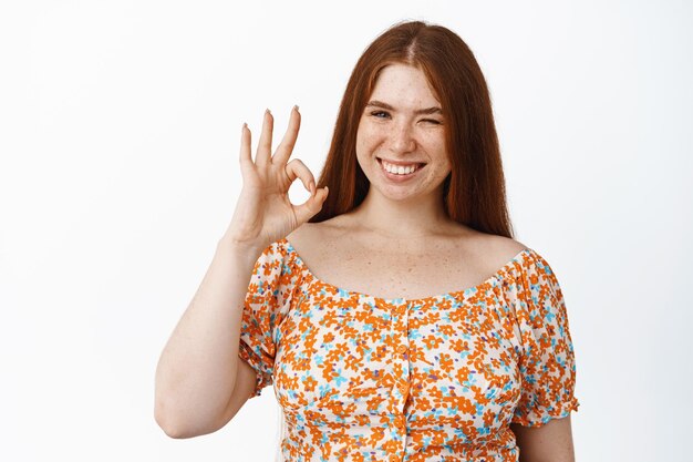 Una alegre chica pelirroja guiñando un ojo y sonriendo mostrando un gesto de acuerdo con una cara satisfecha y confiada recomendando el producto de pie vestido sobre fondo blanco