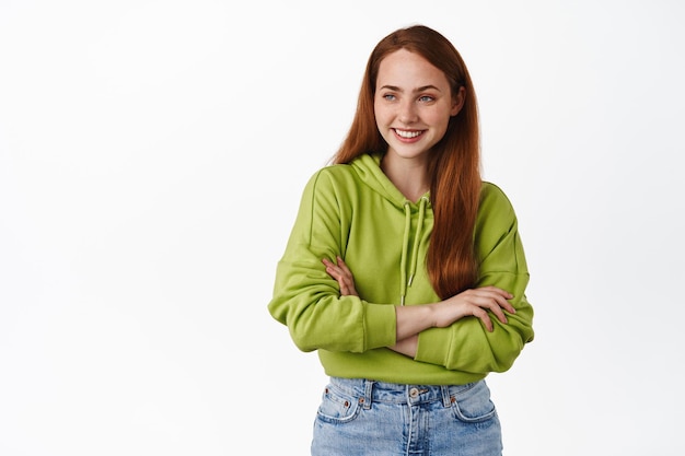 Una alegre chica pelirroja riendo, sonriendo y luciendo despreocupada a un lado, de pie relajada con los brazos cruzados, con capucha verde y jeans, fondo blanco.