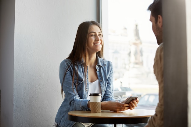 Alegre chica de pelo oscuro en ropa elegante sentado en la cafetería, tomando café, riendo y hablando con un amigo sobre el trabajo. Concepto de estilo de vida.