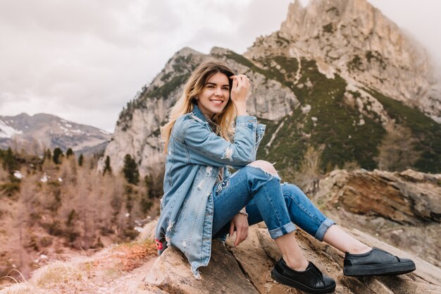 Alegre chica rubia descansa sobre piedra después de escalar las montañas y reír