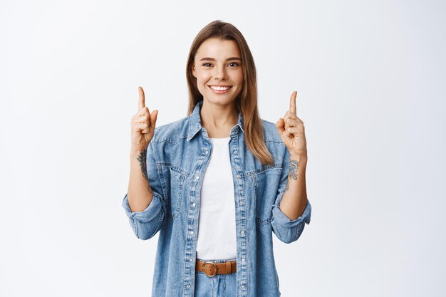 Alegre chica rubia con sonrisa blanca y saludable, apuntando con el dedo hacia el espacio vacío en la parte superior, de pie en blanco y publicidad