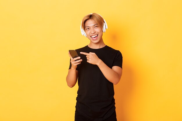 Alegre chico guapo asiático satisfecho, escuchando música o un buen podcast en auriculares, señalando con el dedo al teléfono inteligente con sonrisa complacida, pared amarilla