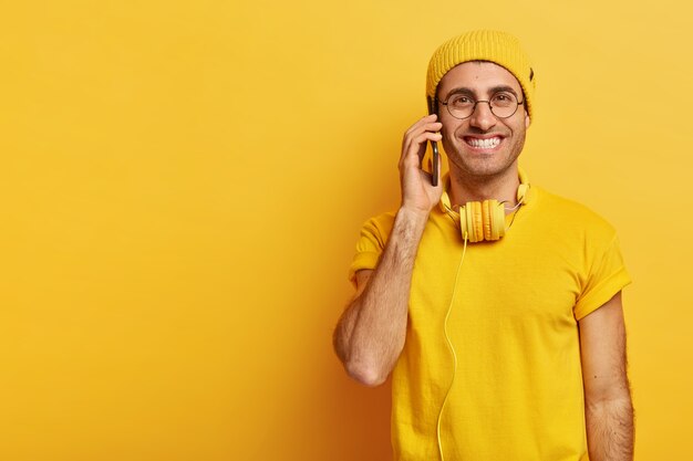 Alegre chico hipster con apariencia agradable, sonríe positivamente, habla a través de un teléfono inteligente, se viste con ropa casual, usa lentes transparentes, habla sobre las últimas tendencias de moda con su mejor amigo