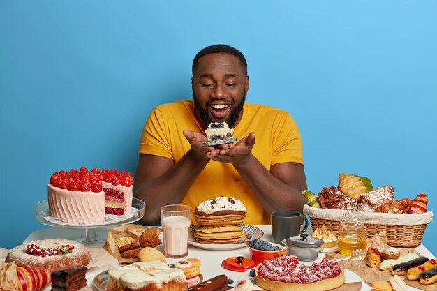 alegre chico de piel oscura, goloso, sostiene un pequeño cupcake, mira felizmente un delicioso postre, viste una camiseta amarilla, posa sobre un fondo azul