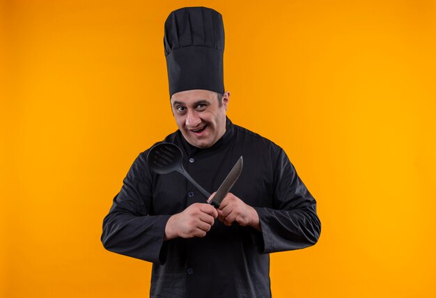 Alegre cocinero de mediana edad en uniforme de chef cruzando el cuchillo y la espátula en la pared amarilla