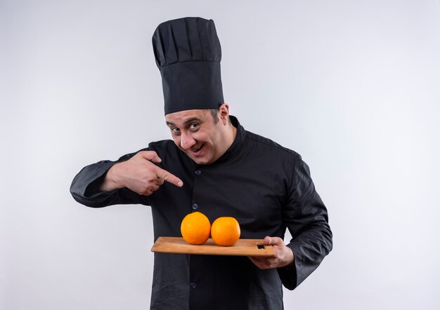 Alegre cocinero de mediana edad en uniforme de chef señala con el dedo a las naranjas en la tabla de cortar en su mano en la pared blanca aislada