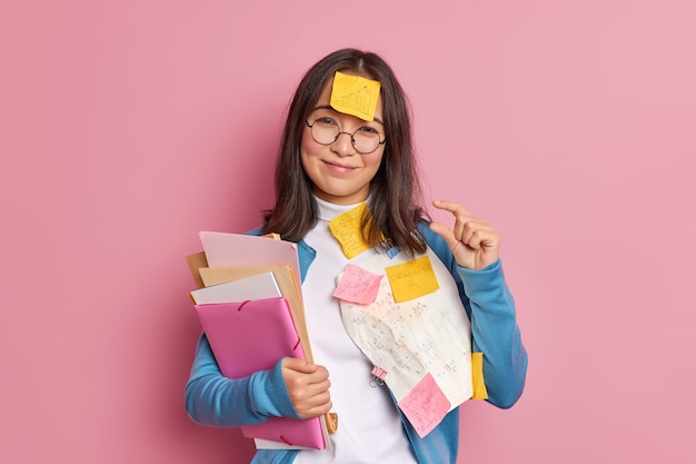 Foto gratuita alegre colegiala morena hace un pequeño gesto que le dice que necesita un poco más de tiempo para la preparación del examen, tiene una nota adhesiva pegada en la frente y tiene fecha límite para terminar el trabajo del proyecto.