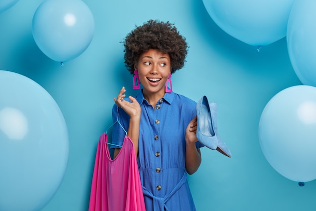 Alegre dama afroamericana positiva elige fuera para la cita, sostiene zapatos azules de tacón alto y vestido rosado en perchas, se prepara para la fiesta y celebración, posa sobre una pared azul con globos inflados