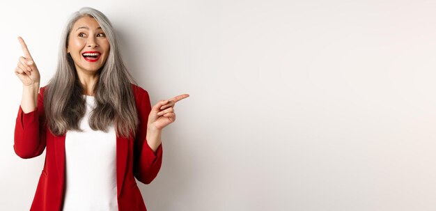 Foto gratuita alegre dama asiática sonriendo señalando con el dedo de lado a dos ofertas promocionales de pie feliz en rojo blaz