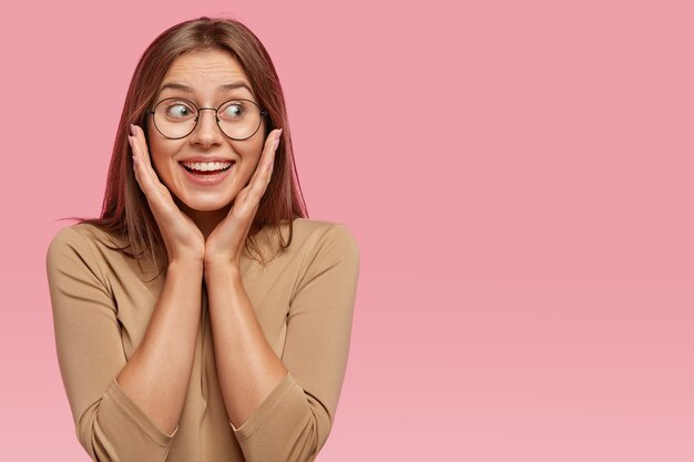 Alegre dama de cabello oscuro con expresión satisfecha, sonrisa agradable en la cara, mantiene las manos en las mejillas