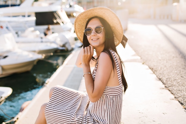 Alegre dama morena con sombrero pasar tiempo en el puerto disfrutando del sol en un día soleado en algún lugar de Europa