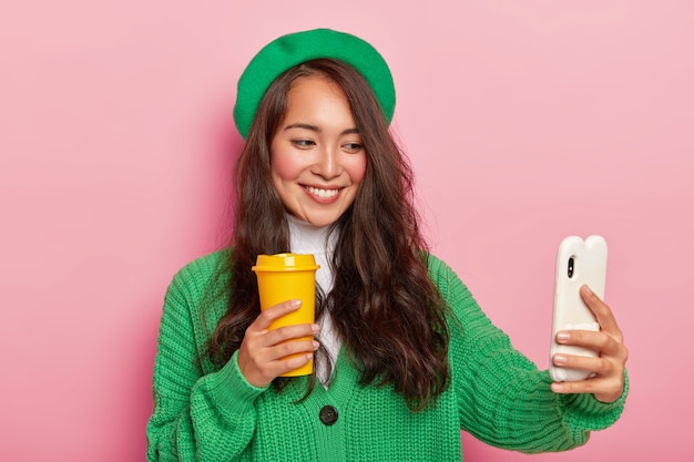 Alegre dama de raza mixta en boina verde y suéter de punto, hace retrato selfie con teléfono celular, posa con taza de café