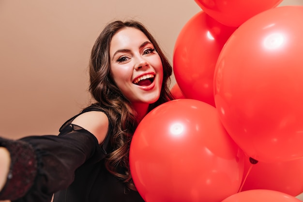 Foto gratuita alegre dama rizada sonríe, mira a cámara y toma selfie con globos.