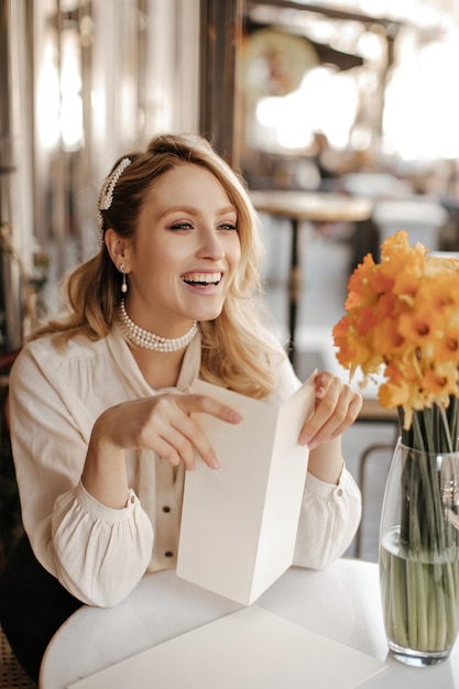 Una alegre dama rubia rizada con una elegante blusa blanca y un collar de perlas sonríe sinceramente sentada en un café de la calle junto a una mesita con un ramo amarillo que sostiene el menú