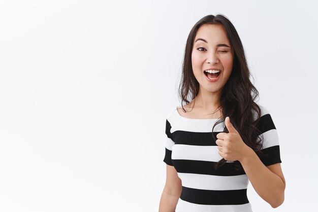 Una alegre y descarada mujer asiática joven con camiseta a rayas muestra el pulgar hacia arriba y guiña un ojo entusiasta insinuando o que le gusta algo juzgando a un amigo increíble vestido nuevo stand fondo blanco complacido